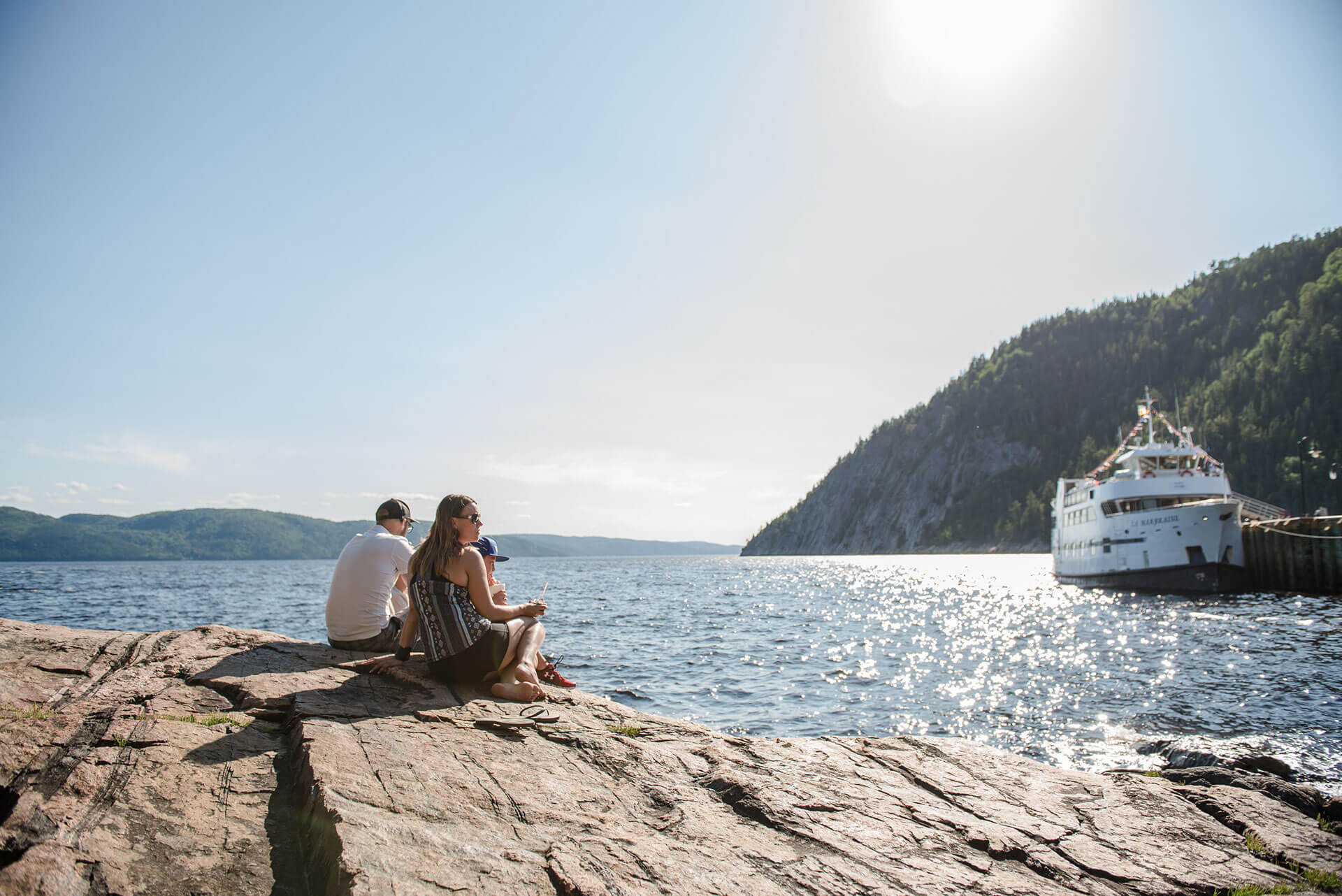 Un père, un mère et leur garçon relaxant sur les roches sur le bord du Fjord du Saguenay admirant le bateau blanc de la croisière La Marjolaine.