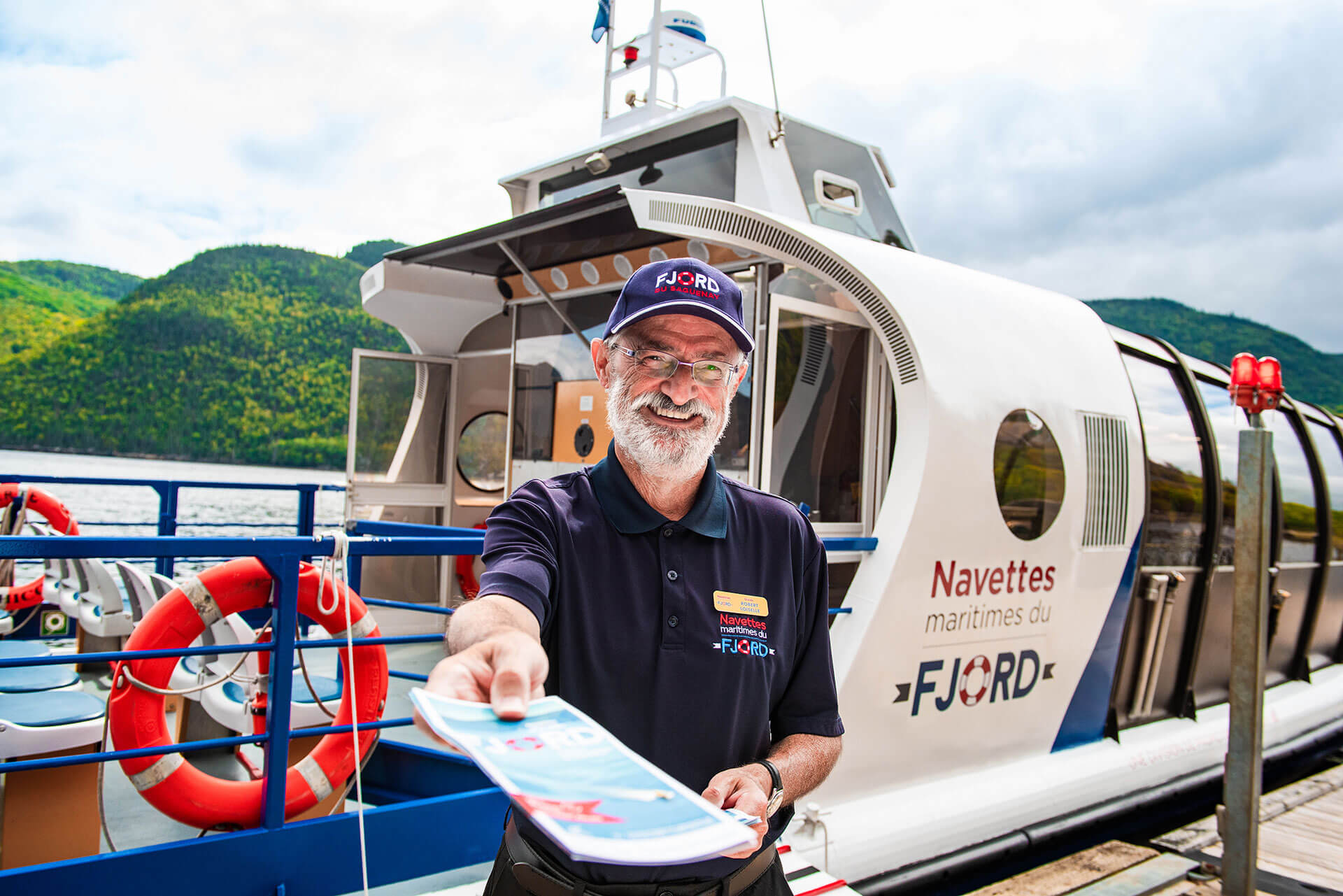 Homme souriant travaillant pour les navettes maritimes du Fjord remettant un dépliant d'information devant un bateau.